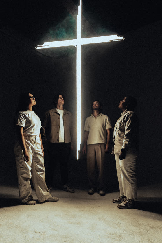 Four people standing beneath a lit cross in a dark room