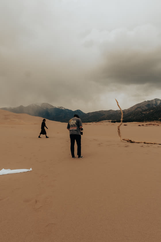 Two figures crossing the sand dunes under a storm sky